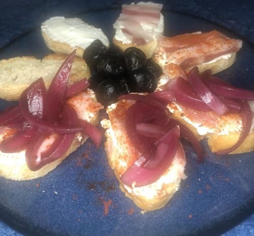 Tapa de rebanada de pan tostado con queso fresco, tocino, pimentón y cebolla morada encurtida