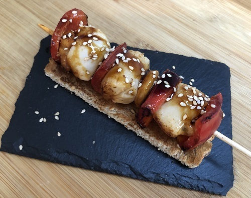 Brocheta de vieiras o zamburiñas con pimientos, salsa teriyaki y sésamo tostado. Sobre pan.